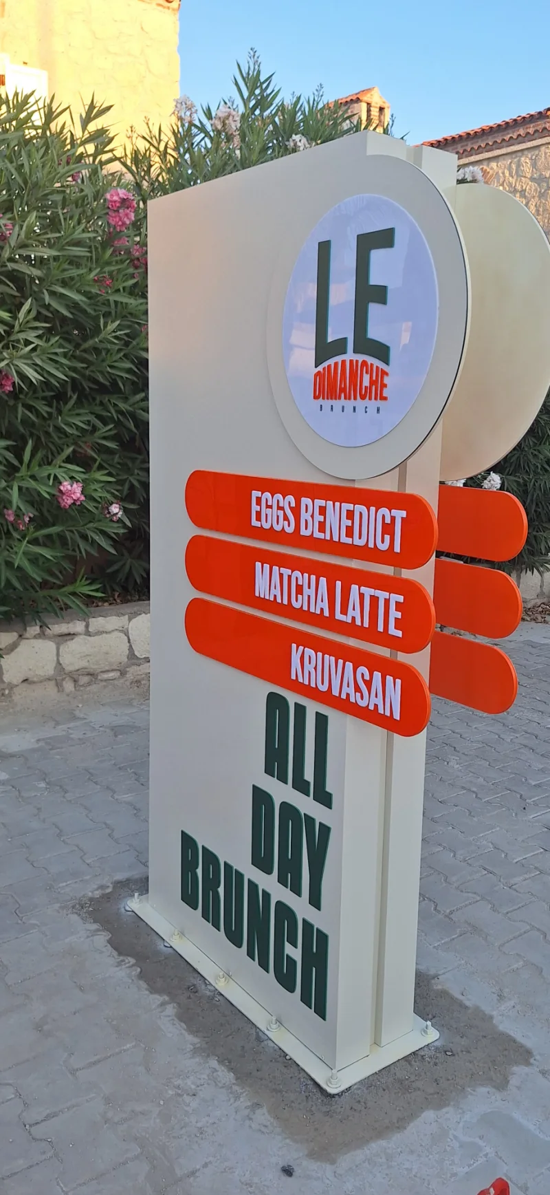 Taş zemine monte edilen dairesel logolu ve turuncu kanatlı dış mekan pilon tabela